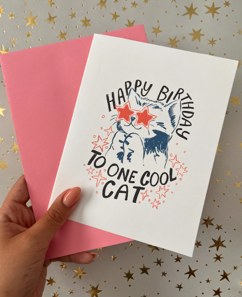 Hand holding a birthday card with a cat illustration and text on a star-patterned background