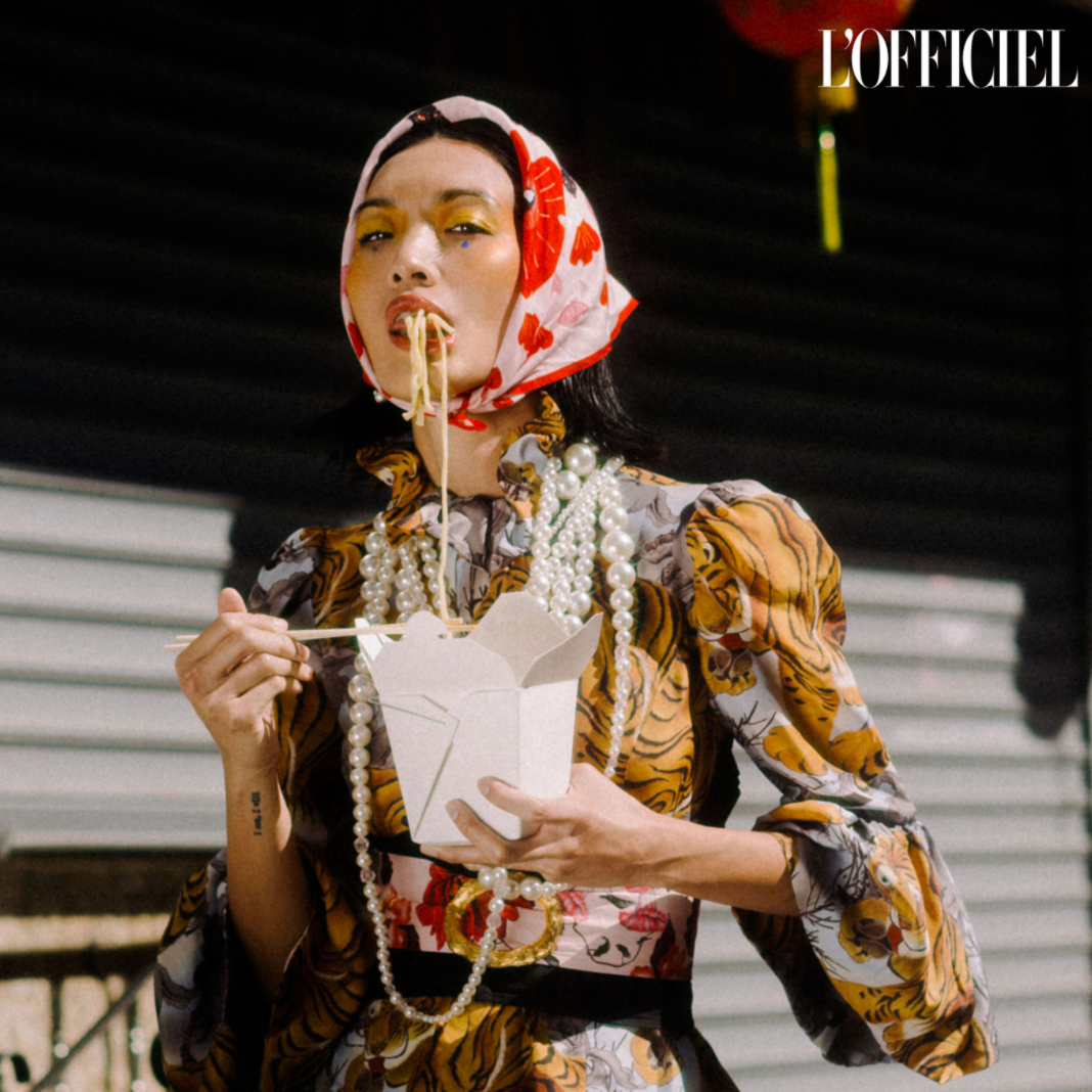 Person eating noodles with a box, wearing a patterned outfit and headscarf, with 'L'Officiel' branding.