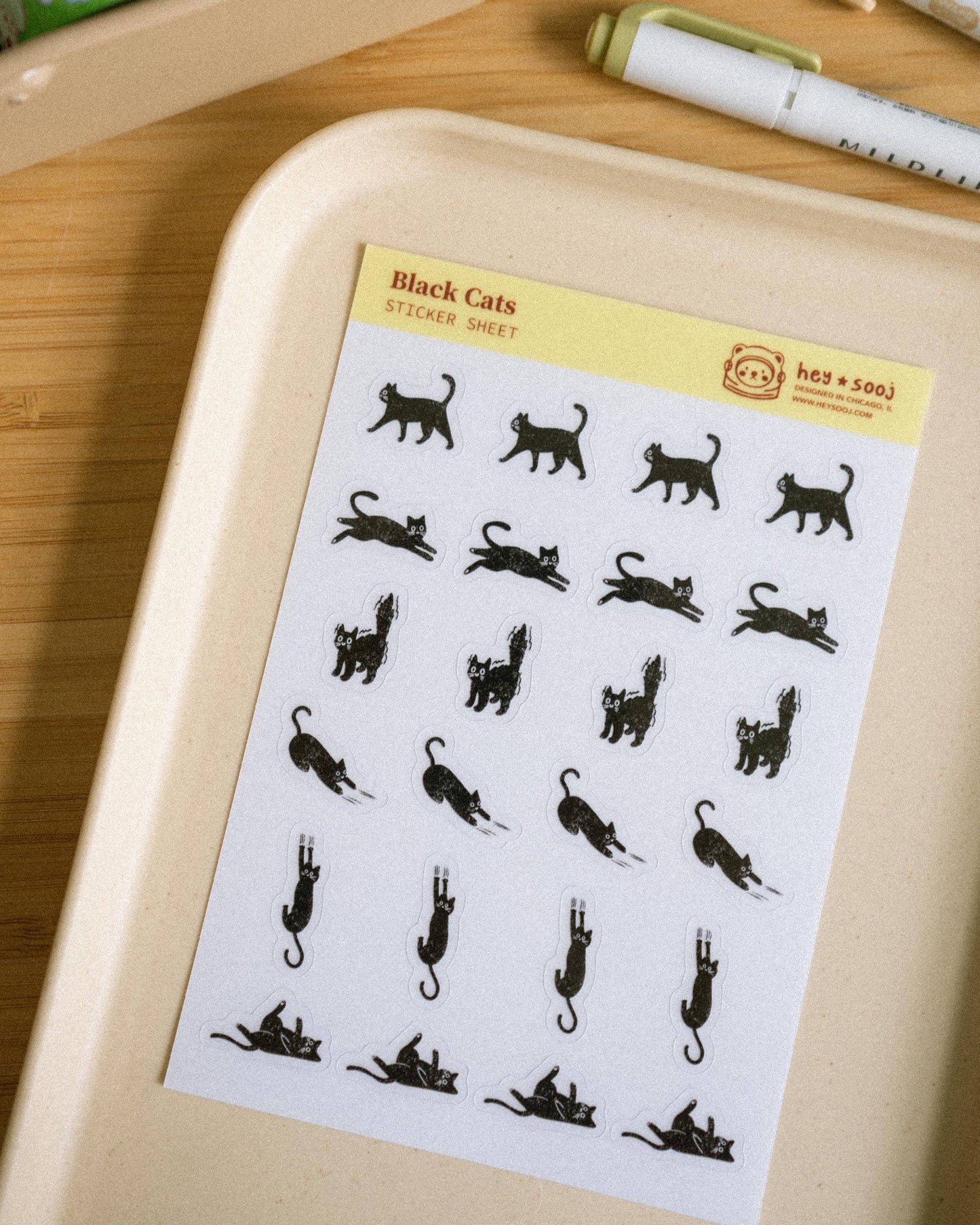 A sheet of black cat silhouette stickers labeled 'Black Cats Sticker Sheet' placed on a beige tray on a wooden desk, with pens and a container visible in the background.