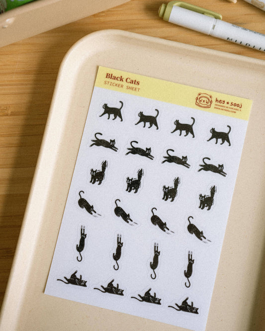 A sheet of black cat silhouette stickers labeled 'Black Cats Sticker Sheet' placed on a beige tray on a wooden desk, with pens and a container visible in the background.