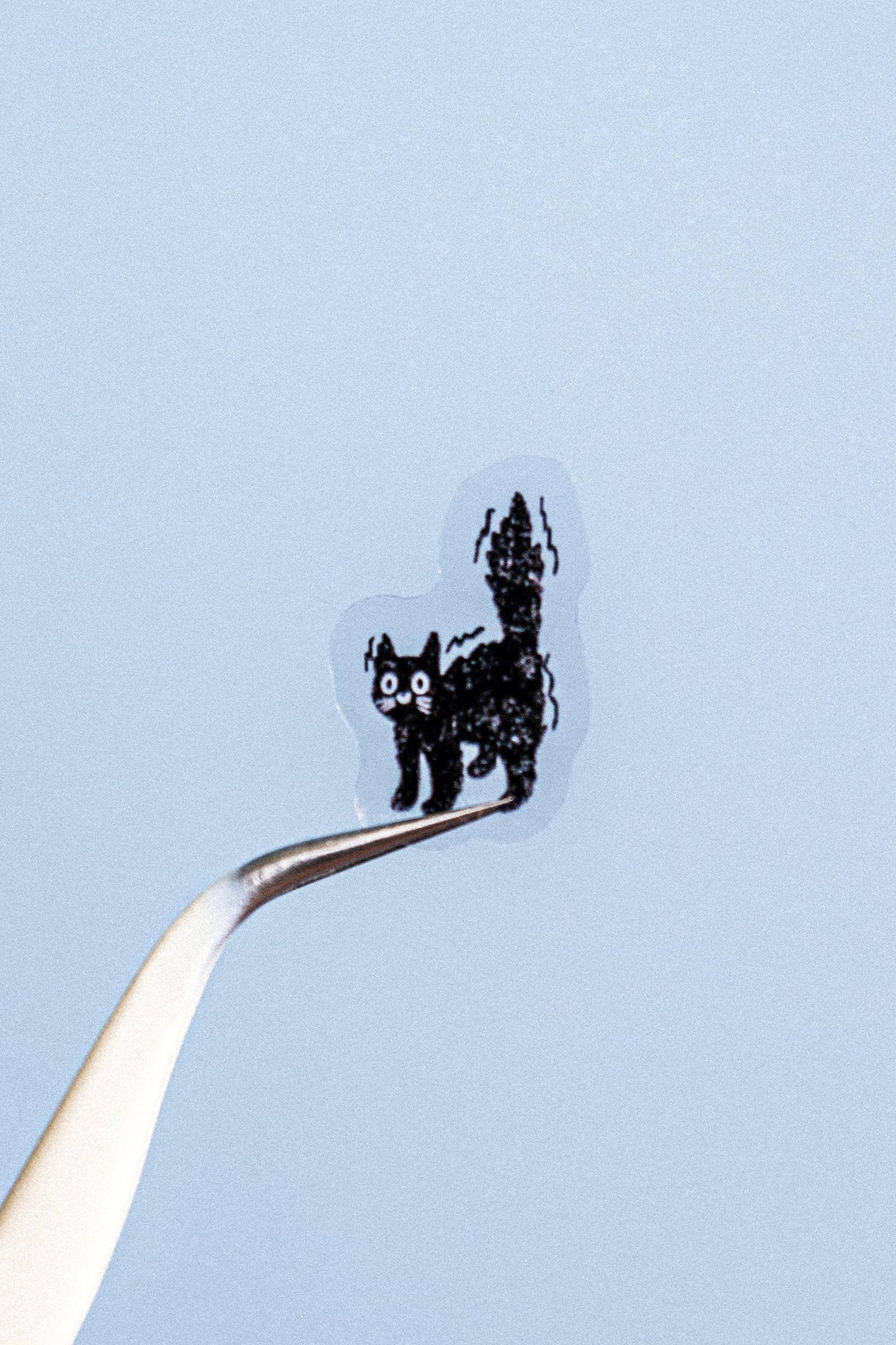A tiny black cat-shaped item with white eyes and a striped tail, held on the tip of a metal tweezer against a pale blue background.