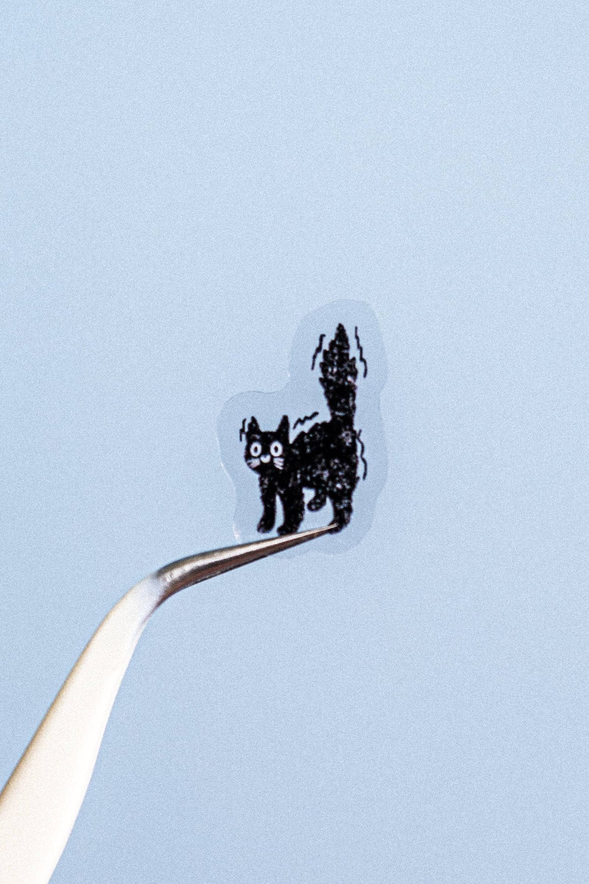 A tiny black cat-shaped item with white eyes and a striped tail, held on the tip of a metal tweezer against a pale blue background.