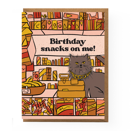 Birthday card with a cat illustration and text 'Birthday snacks on me!' on a colorful bookshelf background.