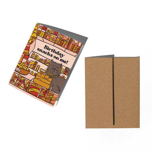 Greeting card with cat illustration and 'Birthday snacks on me!' text, next to a brown envelope.