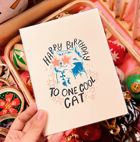 Hand holding a birthday card with a cat illustration and text, surrounded by decorative items.