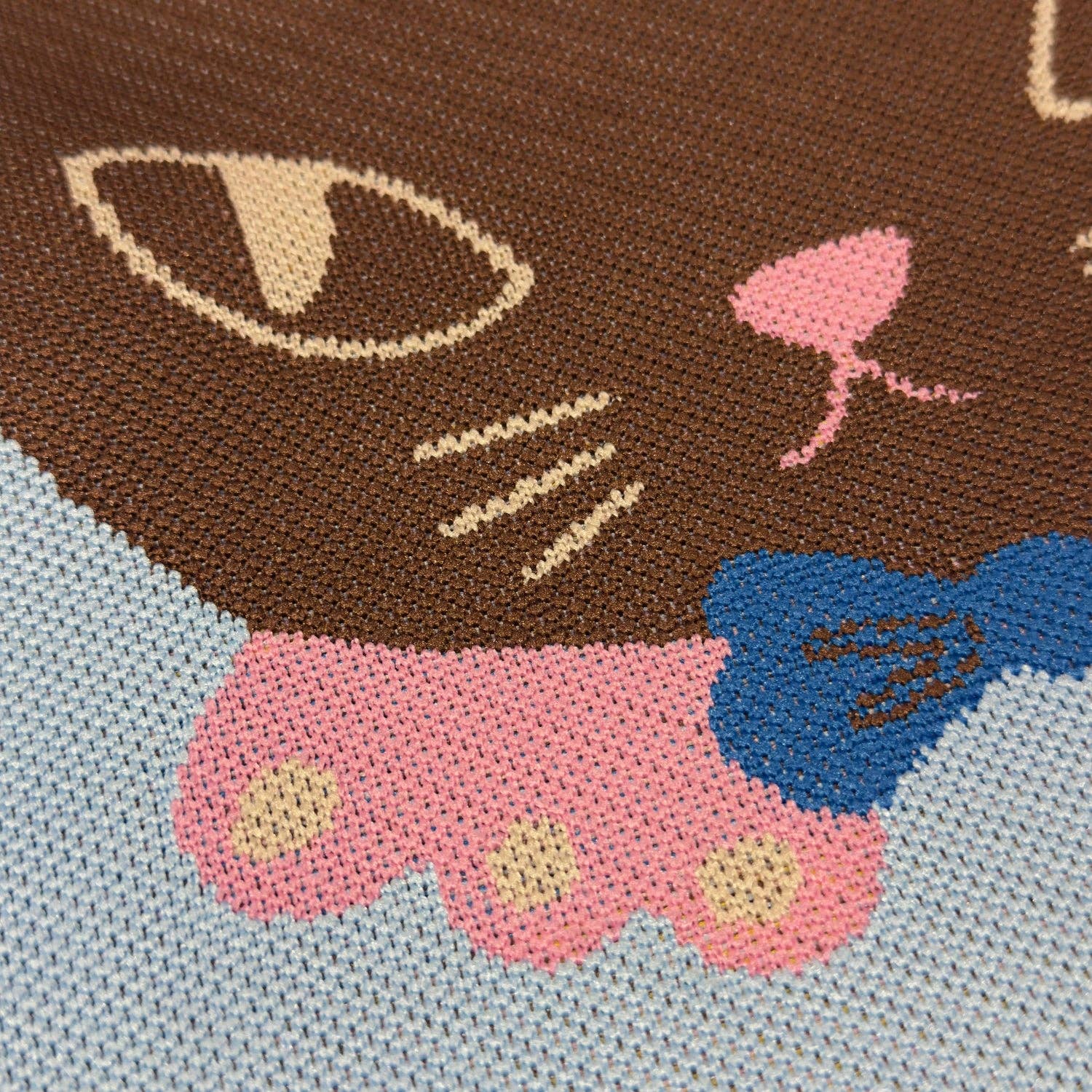 Close-up of a knitted or embroidered design of a cat with a pink bow on a blue background.