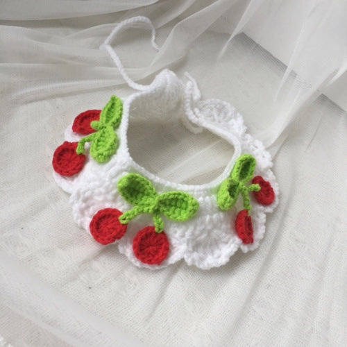 A white crocheted cat collar with a cherry pattern, featuring red crochet cherries and green crochet leaves, laid flat on a white fabric background with sheer white tulle.
