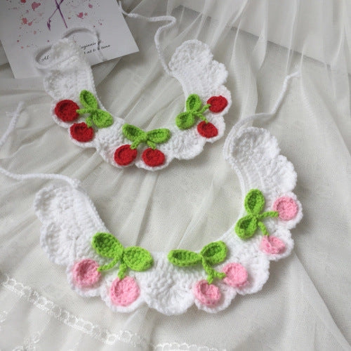 A white crocheted cat collar with a cherry pattern, displayed on a white lace dress. The top version has red cherries with green leaves, and the bottom version has pink cherries with green leaves.