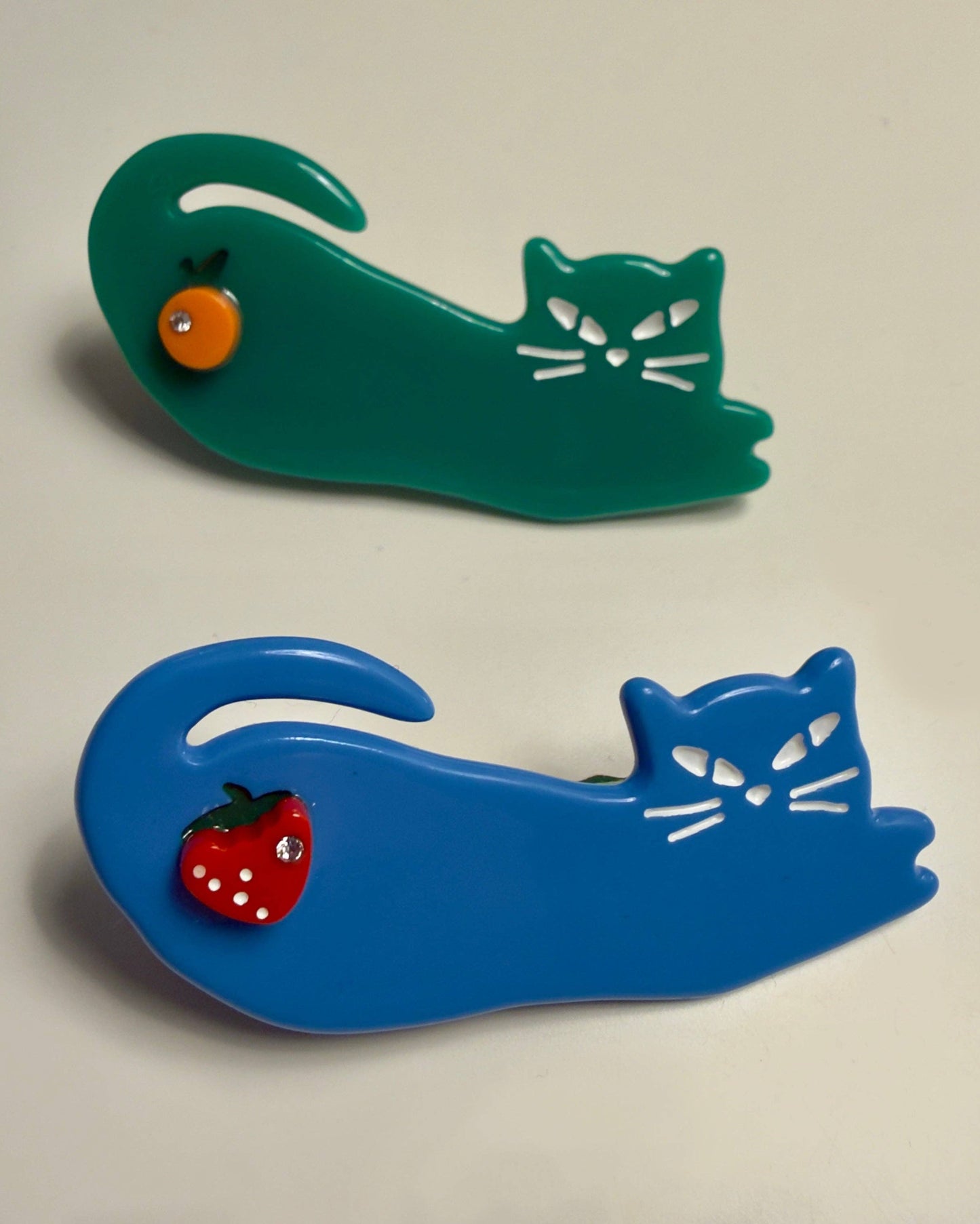 Two cat-shaped brooches, one green and one blue, with small red strawberries on a beige background.