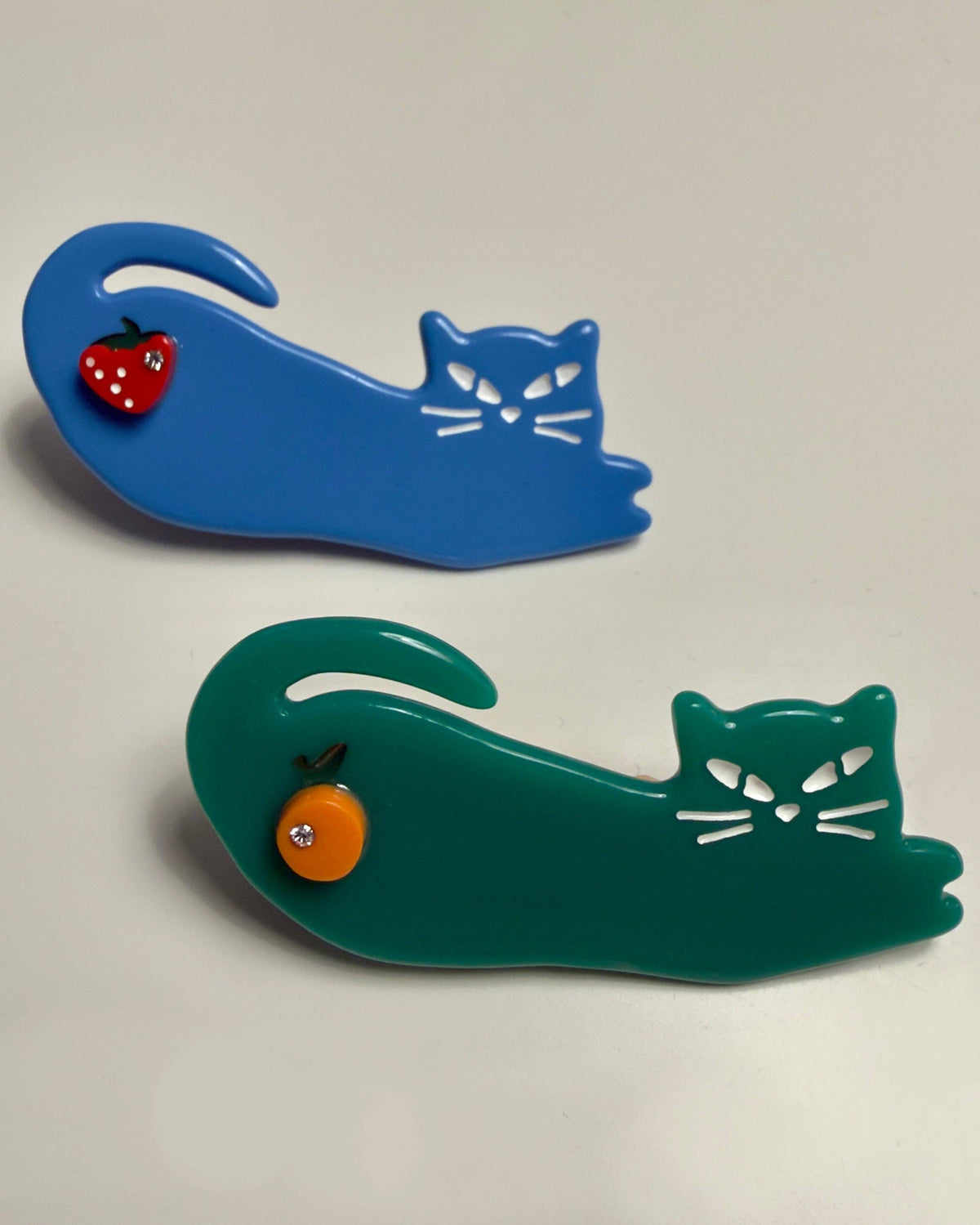 Two cat-shaped hair clips on a light gray background, one in blue with a red strawberry accent and one in green with an orange fruit accent.
