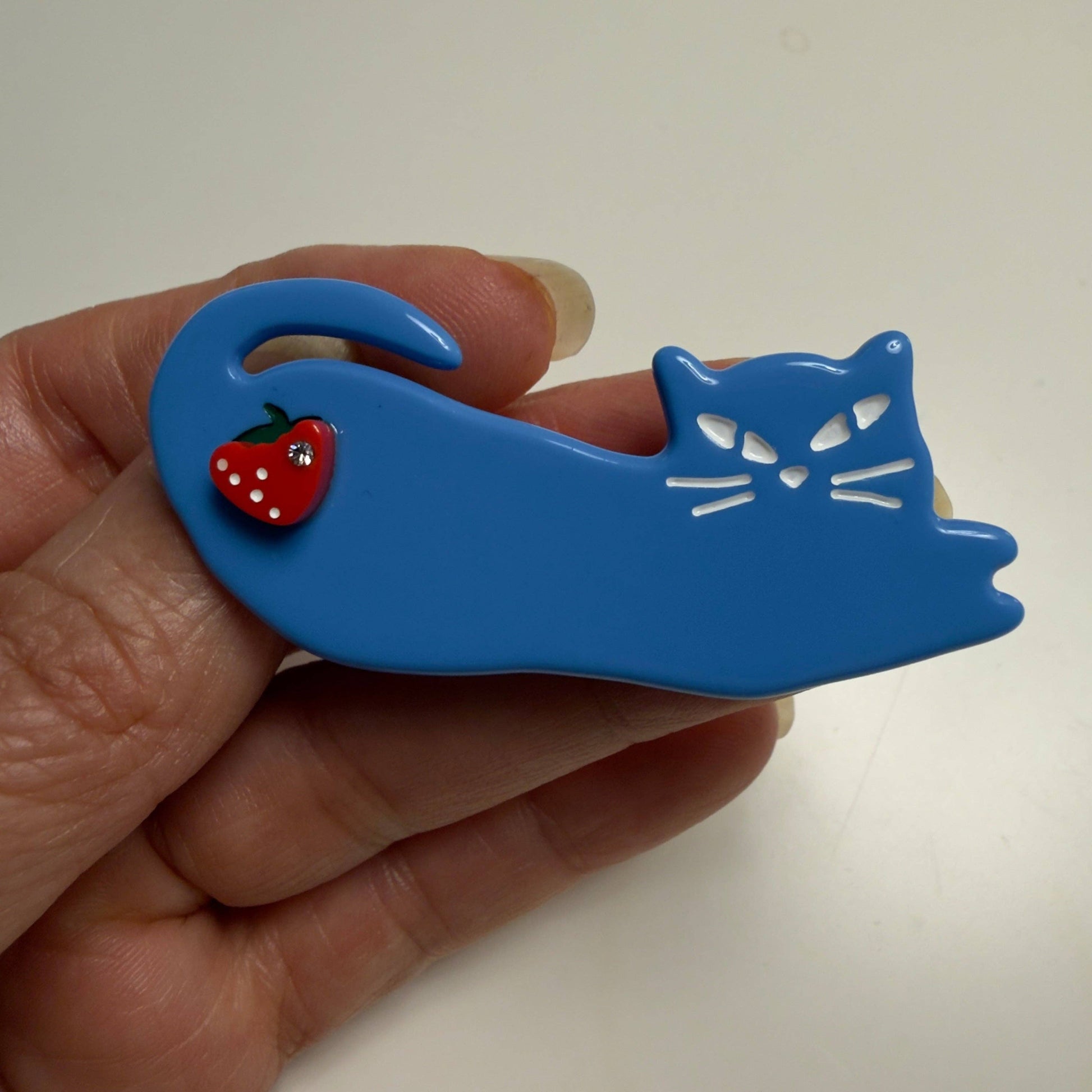 A blue cat-shaped hair claw clip held in a hand, featuring a small red strawberry accent on the side.