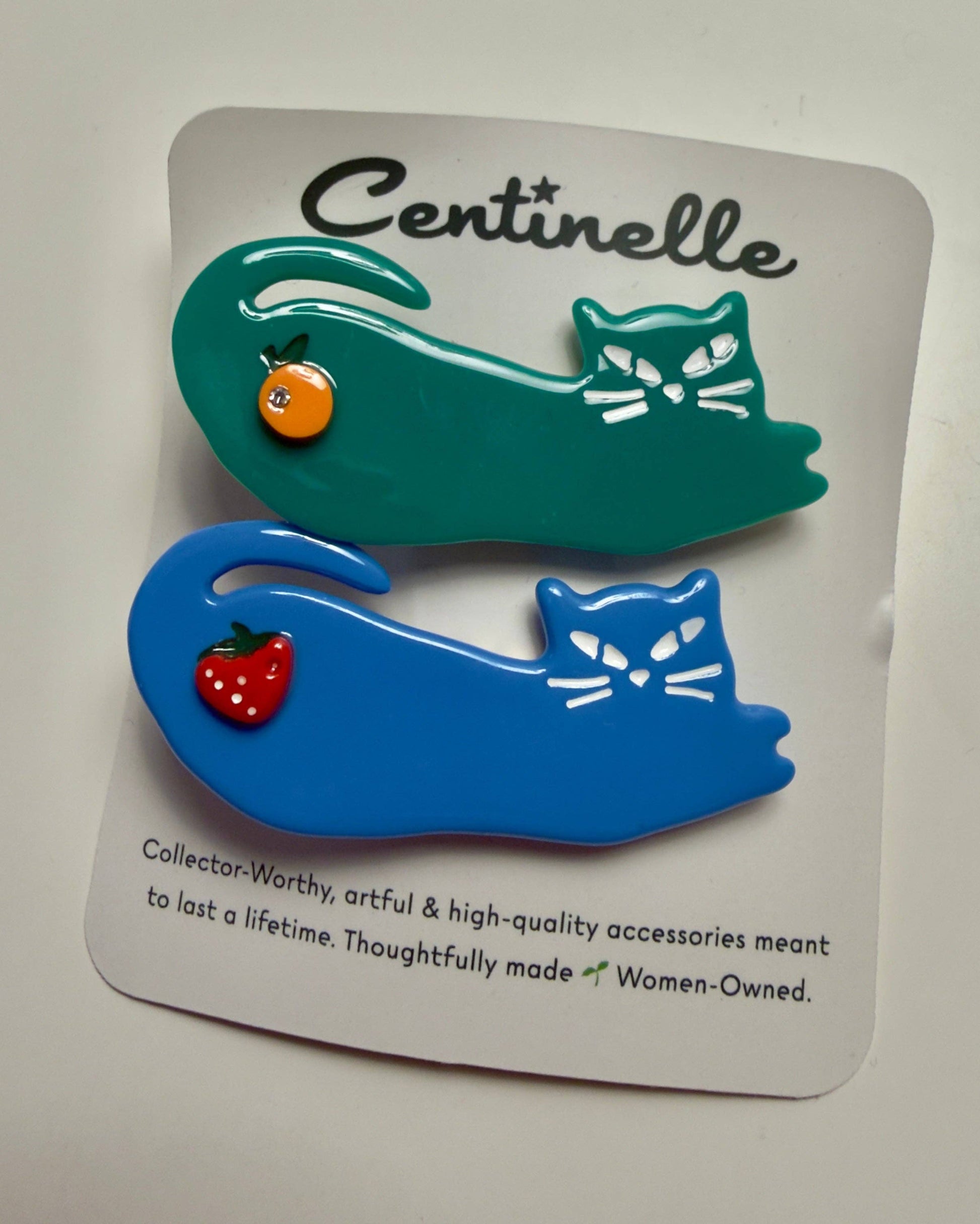 Two colorful cat-shaped hair clips on a branded display card. The top clip is teal with a small orange fruit detail, and the bottom clip is blue with a small red strawberry detail.