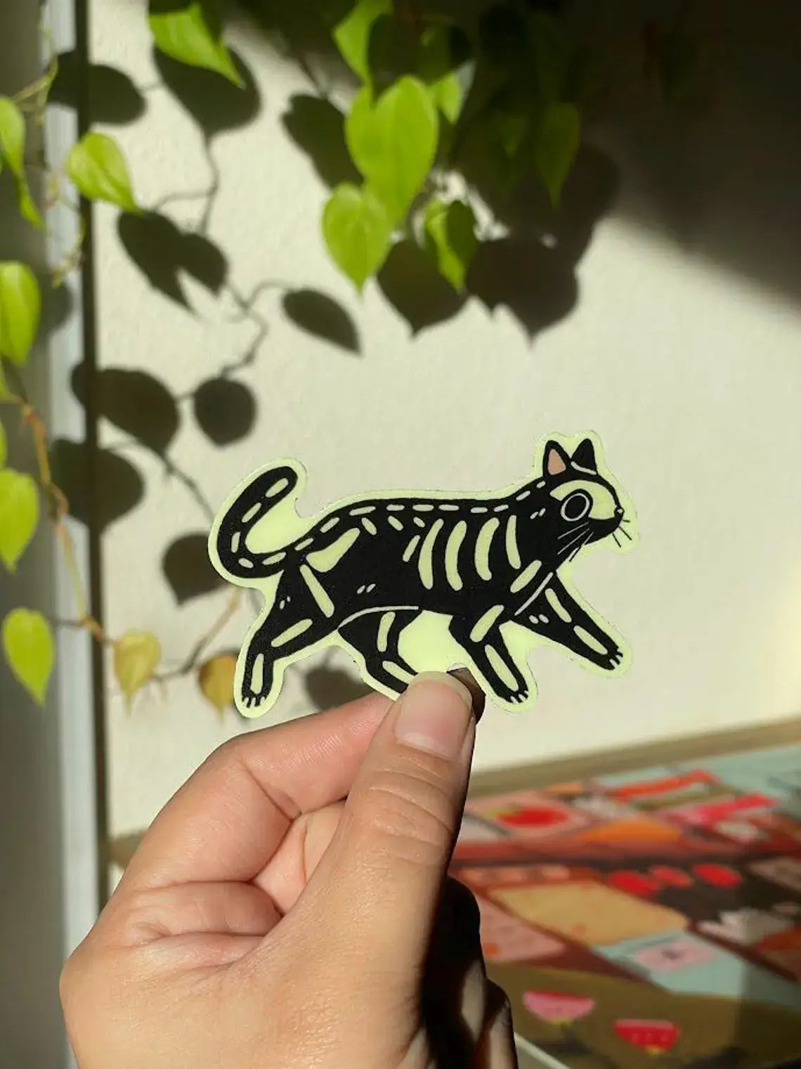 Hand holding a black and white cat illustration against a leafy background