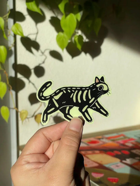 Hand holding a black and white cat illustration against a leafy background