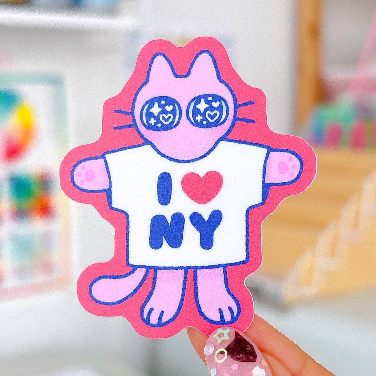 Pink cartoon character with 'I ❤️ NY' shirt held by a hand against a blurred background