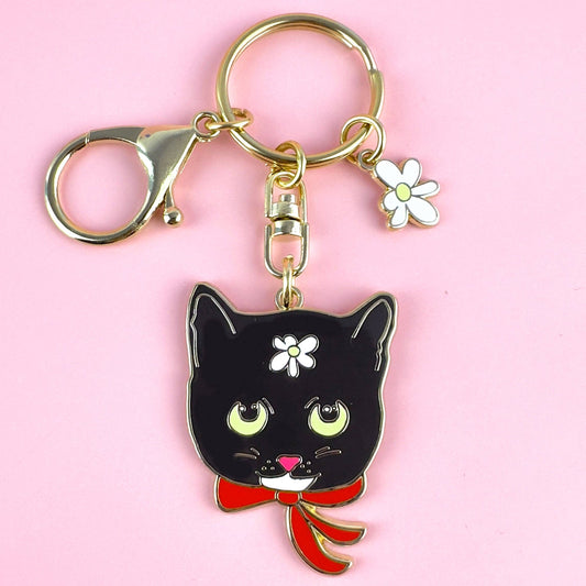 Black cat keychain with a red bow and white flower on a pink background