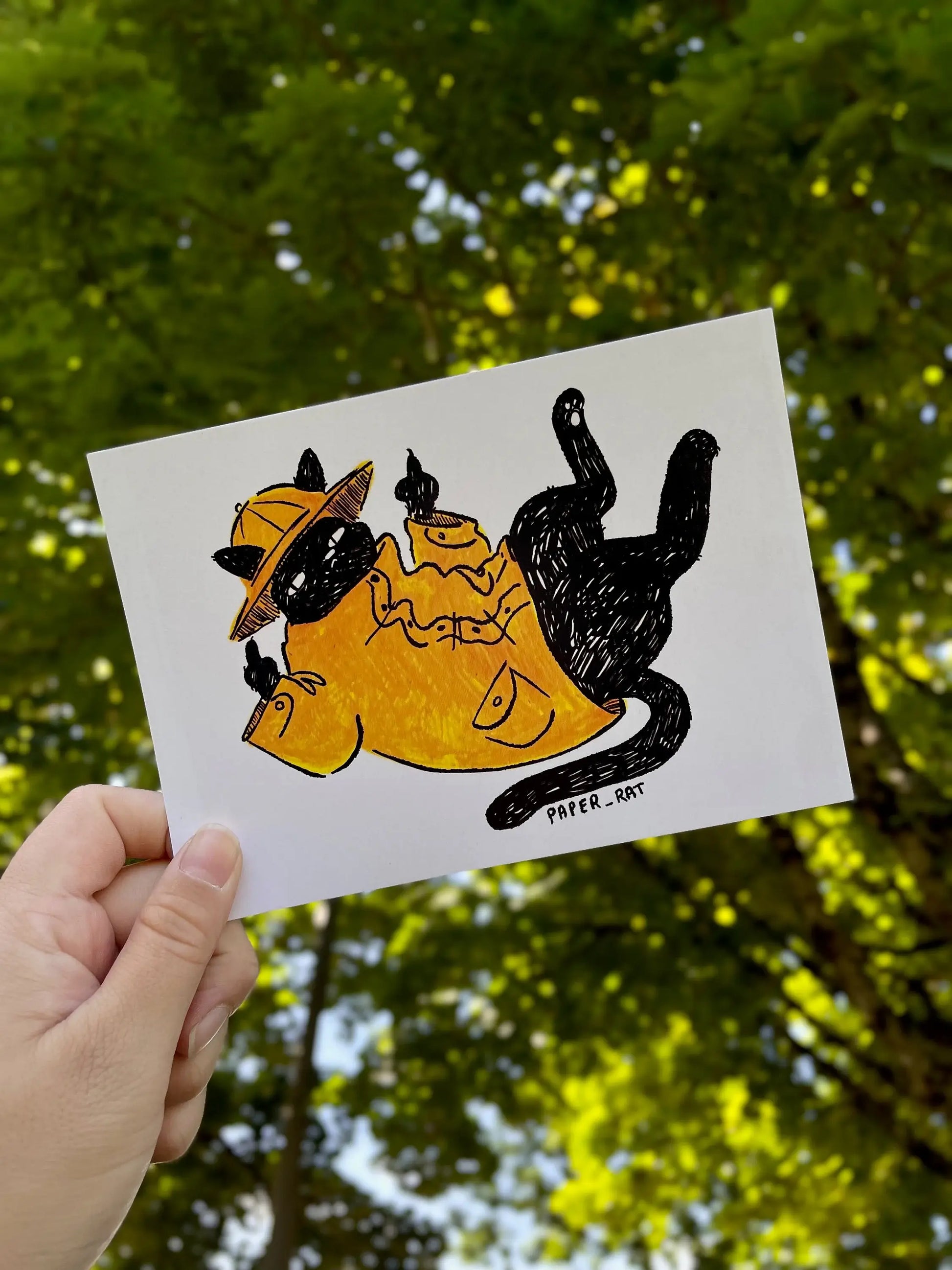 Hand holding a card with a black cat illustration against a green leafy background