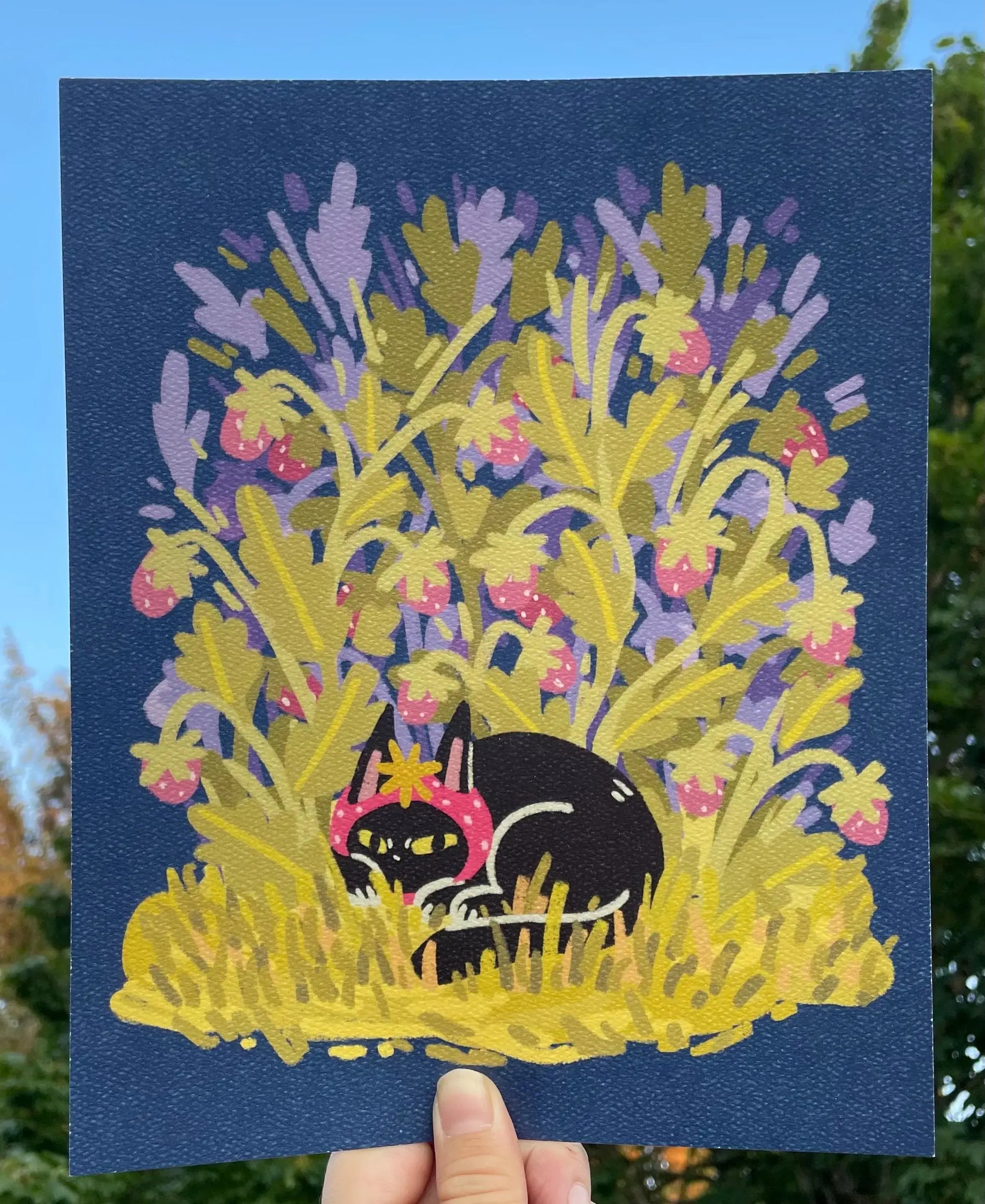 Illustration of a black cat among flowers on a blue card held against a natural background.